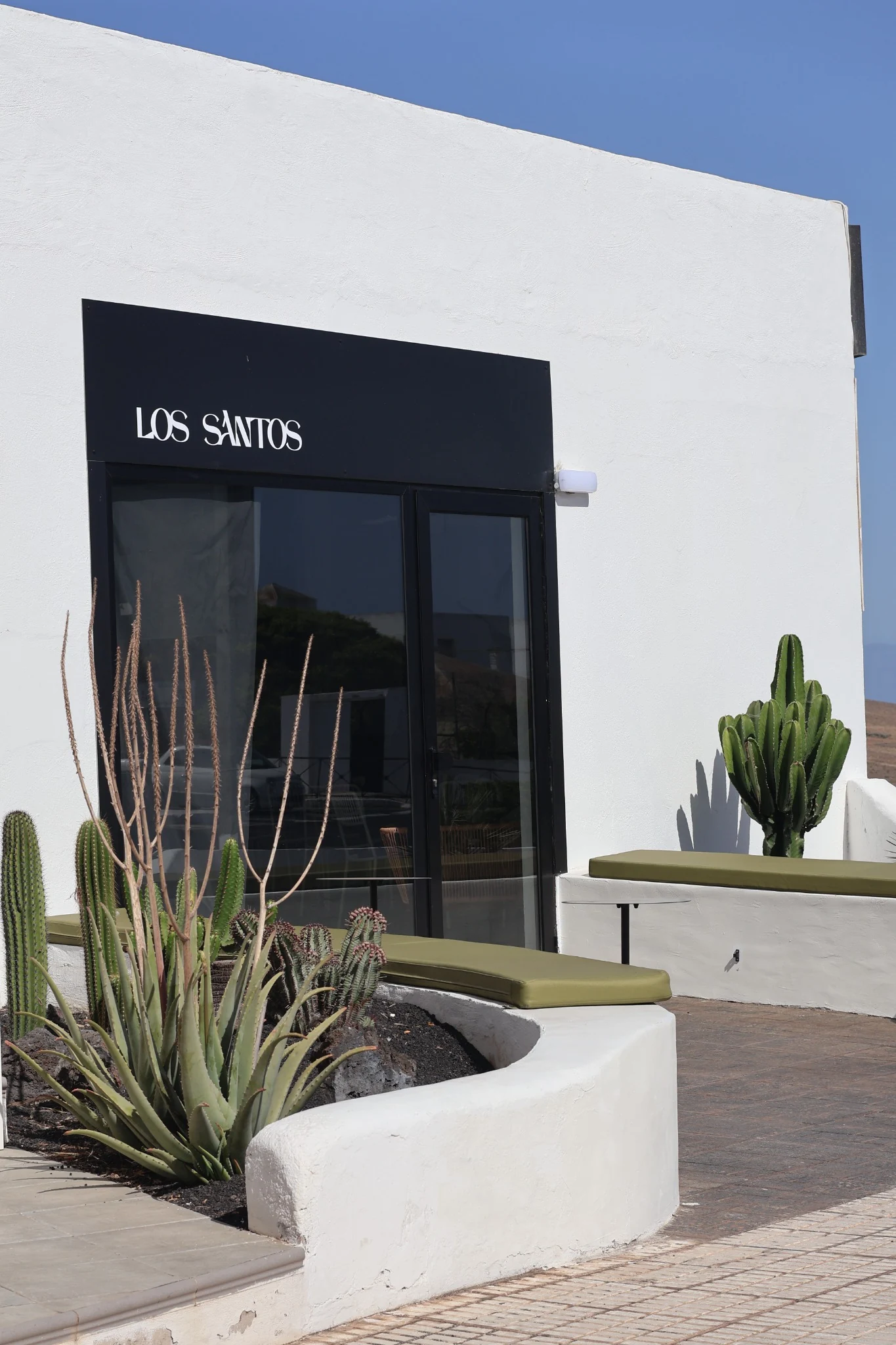 Finished white façade with black Los Santos sign, cacti and olive green seating.